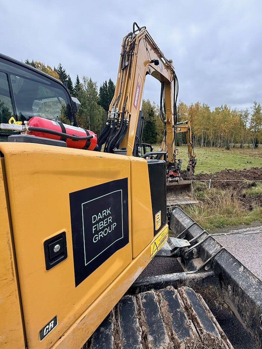 Dark Fiber Group-branded excavator beside a roadside fiber duct trench, with a second excavator digging behind